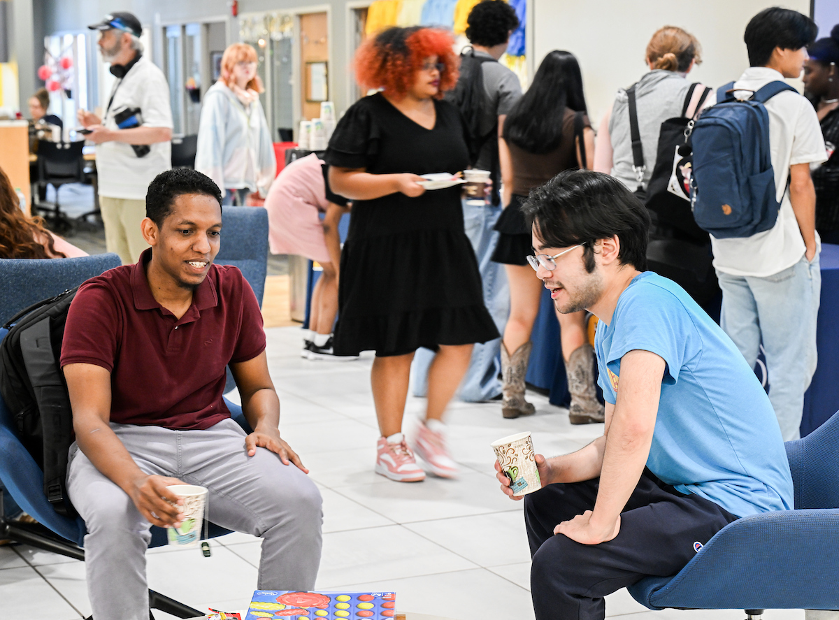 Two students sit in chairs with drinks in hand, talking while a group of students stands in the background.