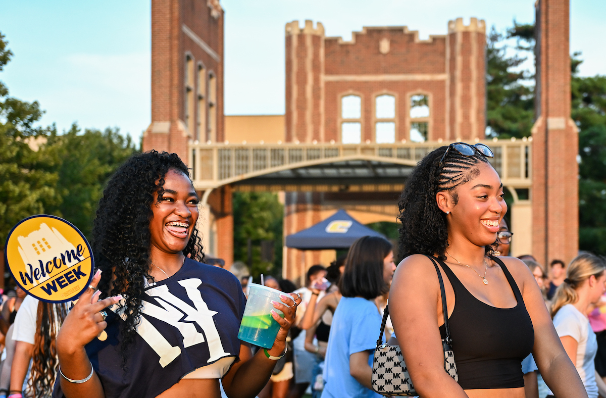 Two students smile and walk at Welcome Week on Chamberlain Field, one holding a lemonade drink.