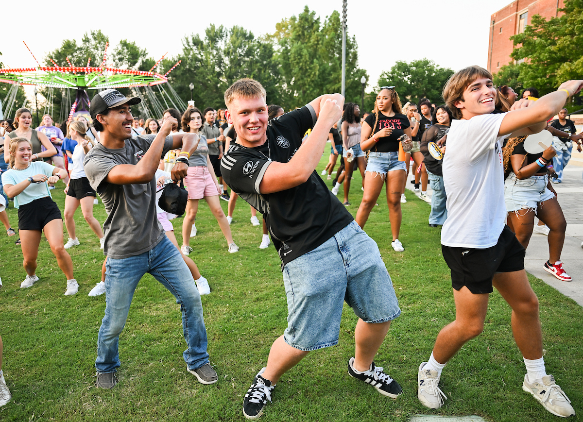 Students dance together on the grass at Welcome Week with a carnival ride in the background.