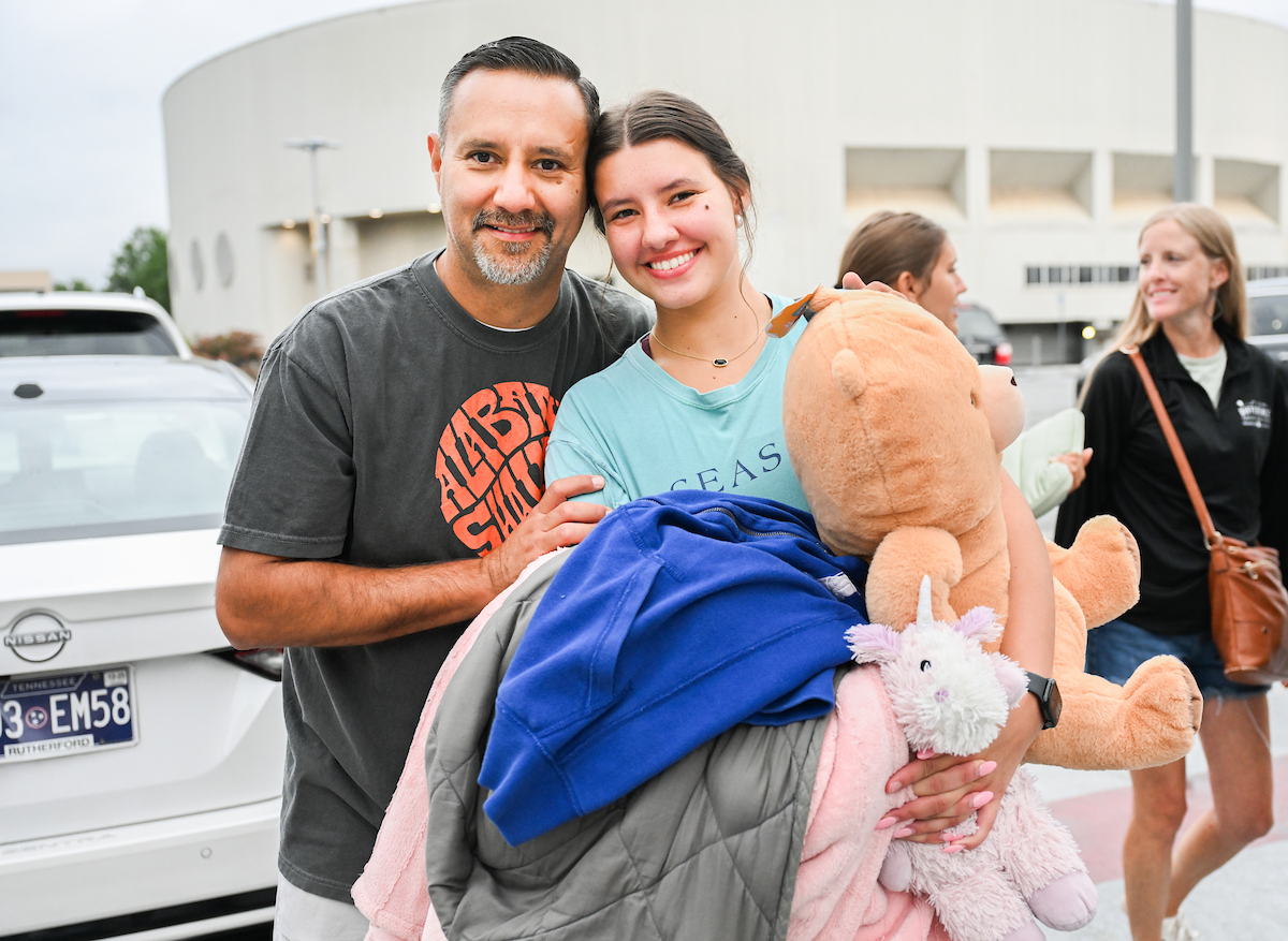 A freshman student holds stuffed animals as she poses next to her father. They are smiling and looking into the camera.
