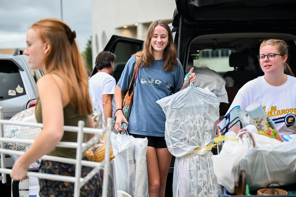 A freshman students holds a hanger with a dress after getting it out of the trunk of a car. She is smiling.