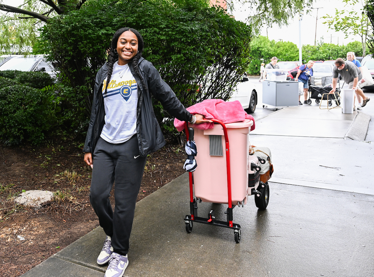 A volunteer at Operation Move in rolls in a students belongings on the sidewalk. She is smiling and looking into the camera.