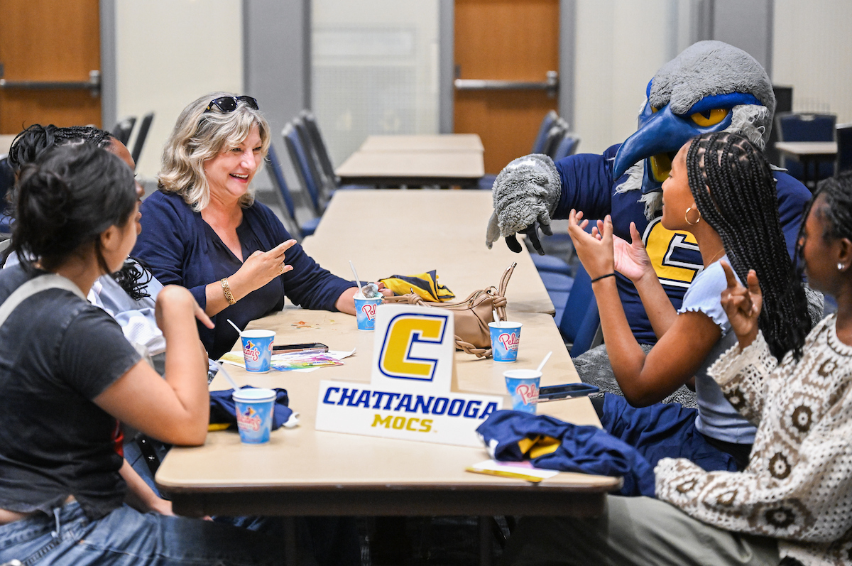 Chancellor Lori Bruce sits across from Scrappy the mascot at the University Center. Students sit around them as she teaches them how to do a "Power C" symbol