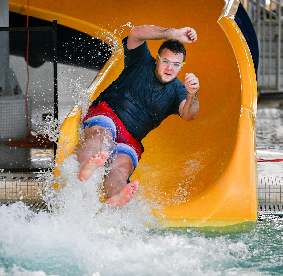 A student wearing goggles and a dark shirt comes down the yellow water slide into the pool.