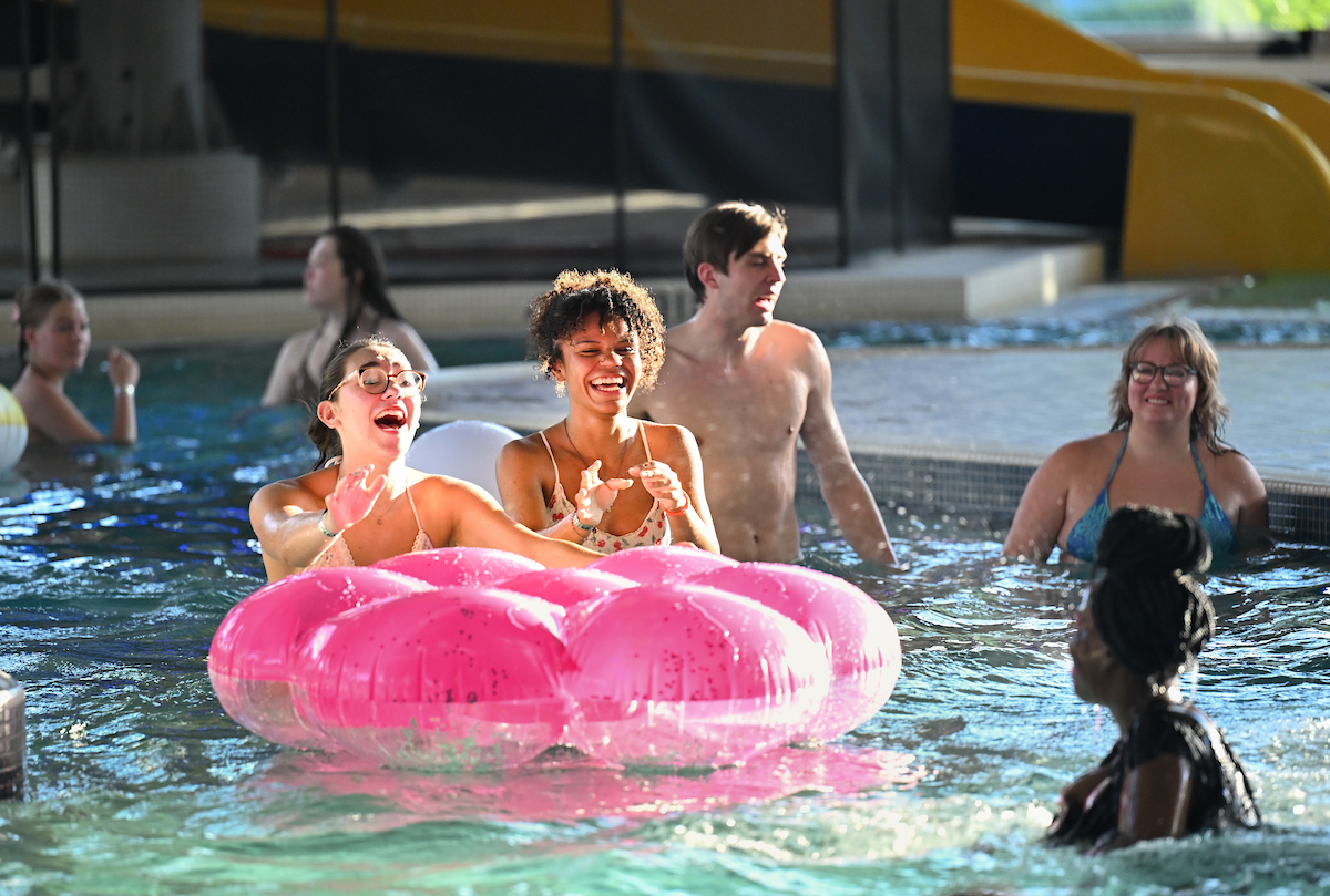 Two students laugh while sitting on a large pink inflatable float in the pool, surrounded by other students swimming.