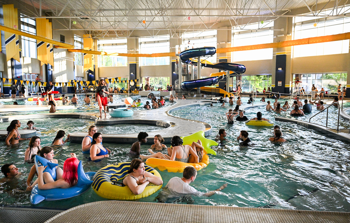A large group of students swim and float on inflatables in the Aquatic and Recreation Center pool during the Soundwaves Pool Party, with the yellow and blue water slide visible in the background.