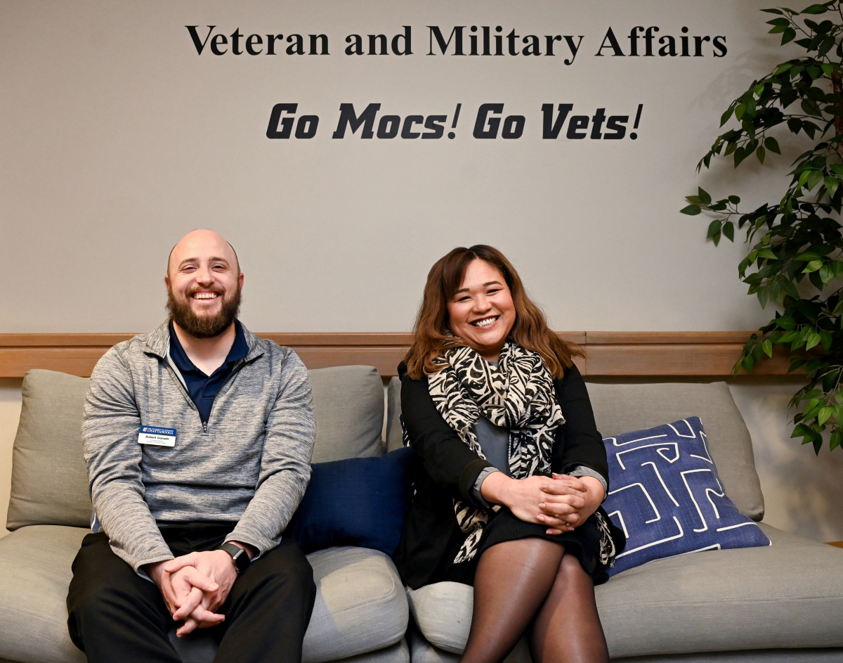 Associate Director Robert Dorsett and Director Sylvana Matthews sitting on a couch under a sign that reads "Veteran and Military Affairs. Go Mocs! Go Vets!"
