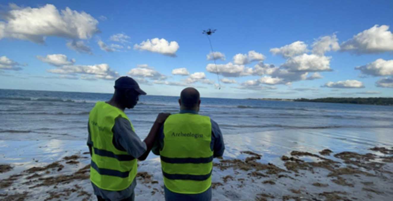 Two men look out to sea from a beach with a drone flying overhead.