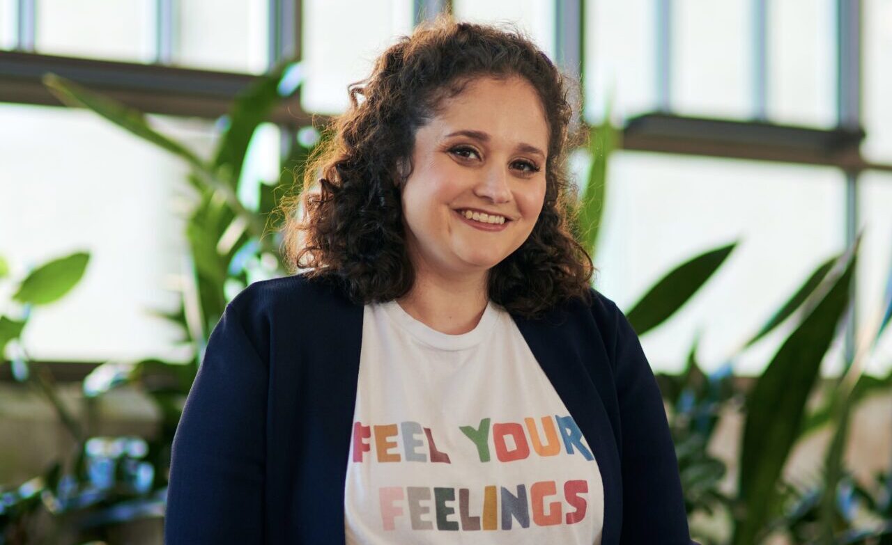 A smiling person with curly brown hair sits indoors in front of large windows and green plants. They are wearing a navy blazer over a white T-shirt with colorful block letters that read “FEEL YOUR FEELINGS.”