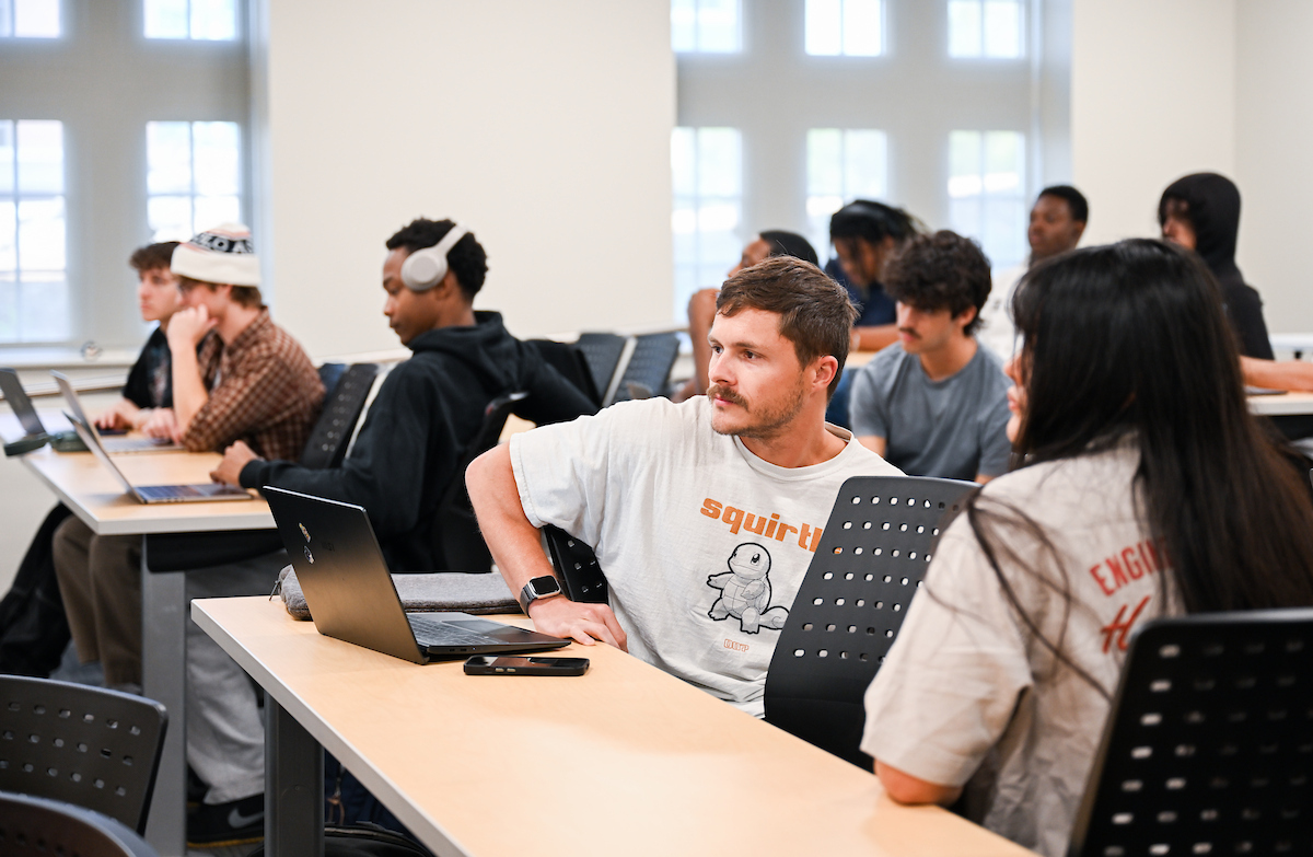 Cody Posey sits in a classroom with classmates. He sits in front of an open laptop and is faced toward the front.