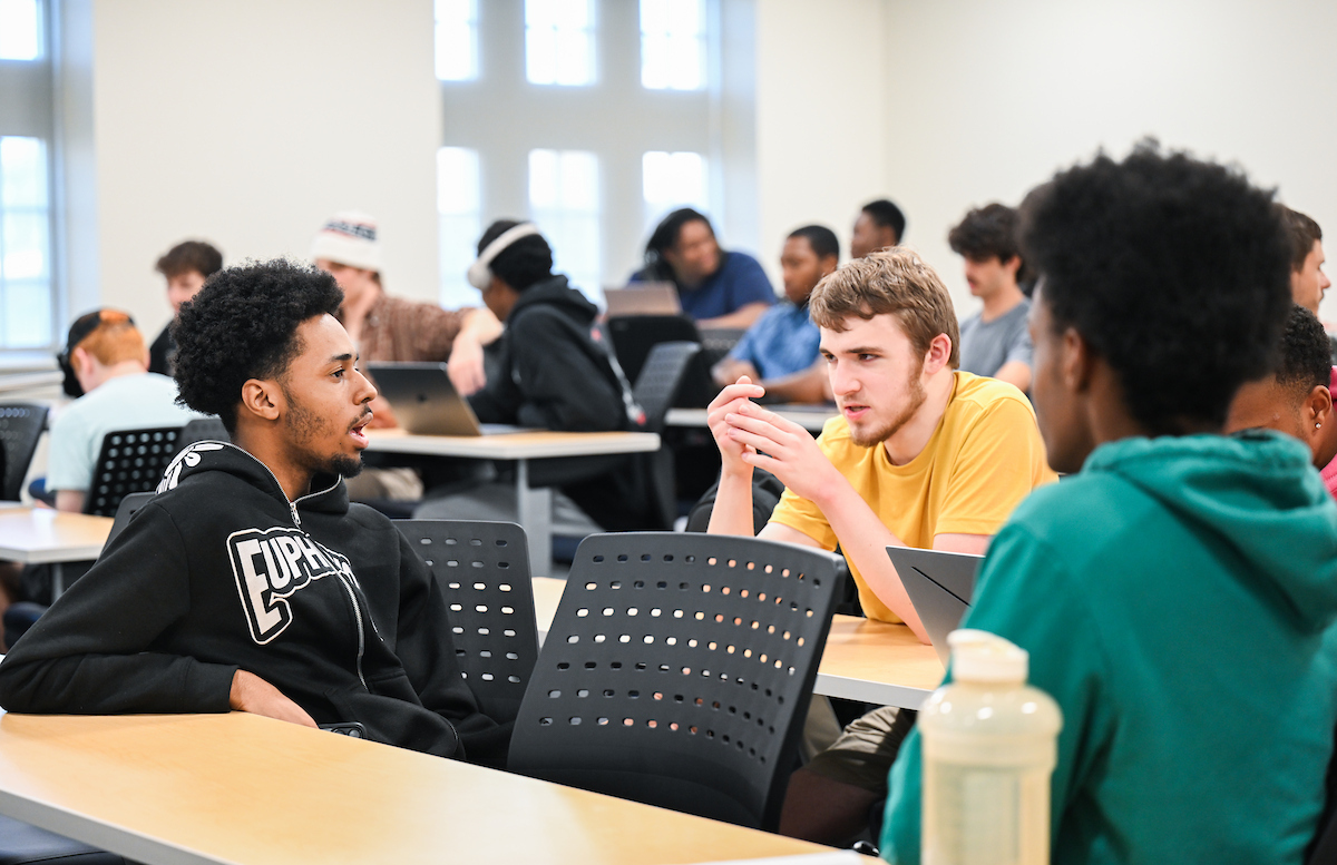 Three students sitting in a full classroom talk with each other.