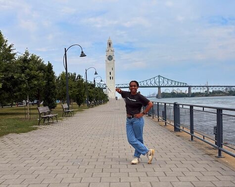 Student Hailey Henderson poses playfully on a riverside walkway, with a distant clock tower and bridge behind her.