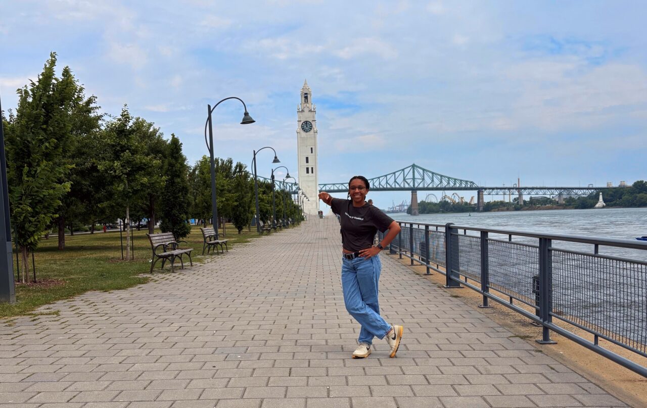 A person stands smiling on a wide paved walkway along a river, pointing toward a tall white clock tower in the distance. Trees and benches line one side of the path, while black street lamps and a metal railing run along the river’s edge. In the background, a large green truss bridge spans the river under a partly cloudy sky.