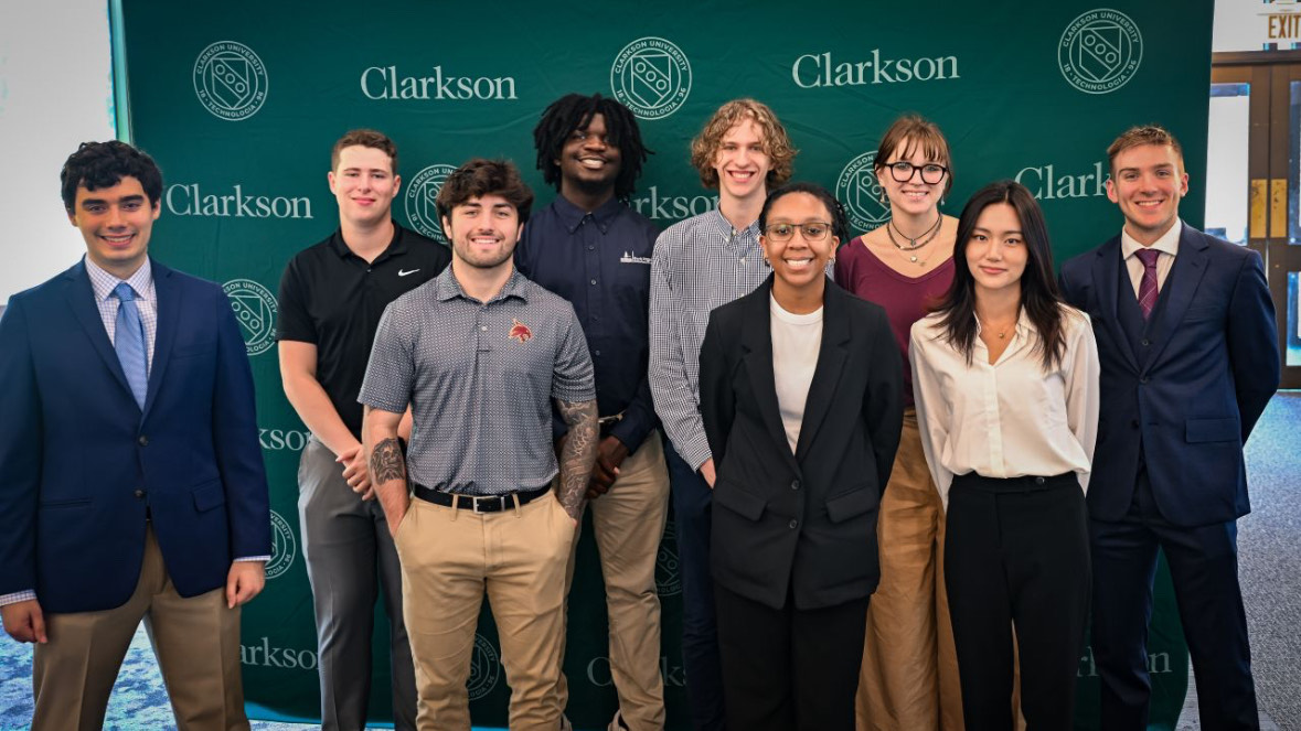 A group of nine students poses in front of a green Clarkson University backdrop. They stand in two rows, smiling at the camera. The students are dressed in a mix of business and business casual attire — some in suits and ties, others in collared shirts and slacks. The backdrop features the Clarkson University name and seal in white.