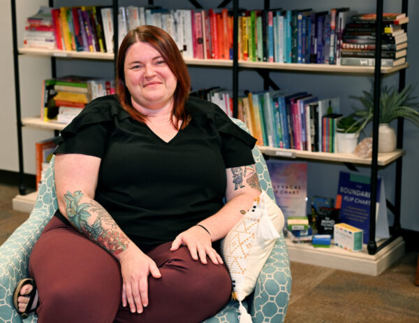 A person with red hair wearing a black blouse and burgundy pants sits smiling in a teal-patterned chair. Their arms, which feature colorful floral tattoos, rest on their lap. Behind them is a tall bookshelf filled with books arranged by color, plus small plants and décor.