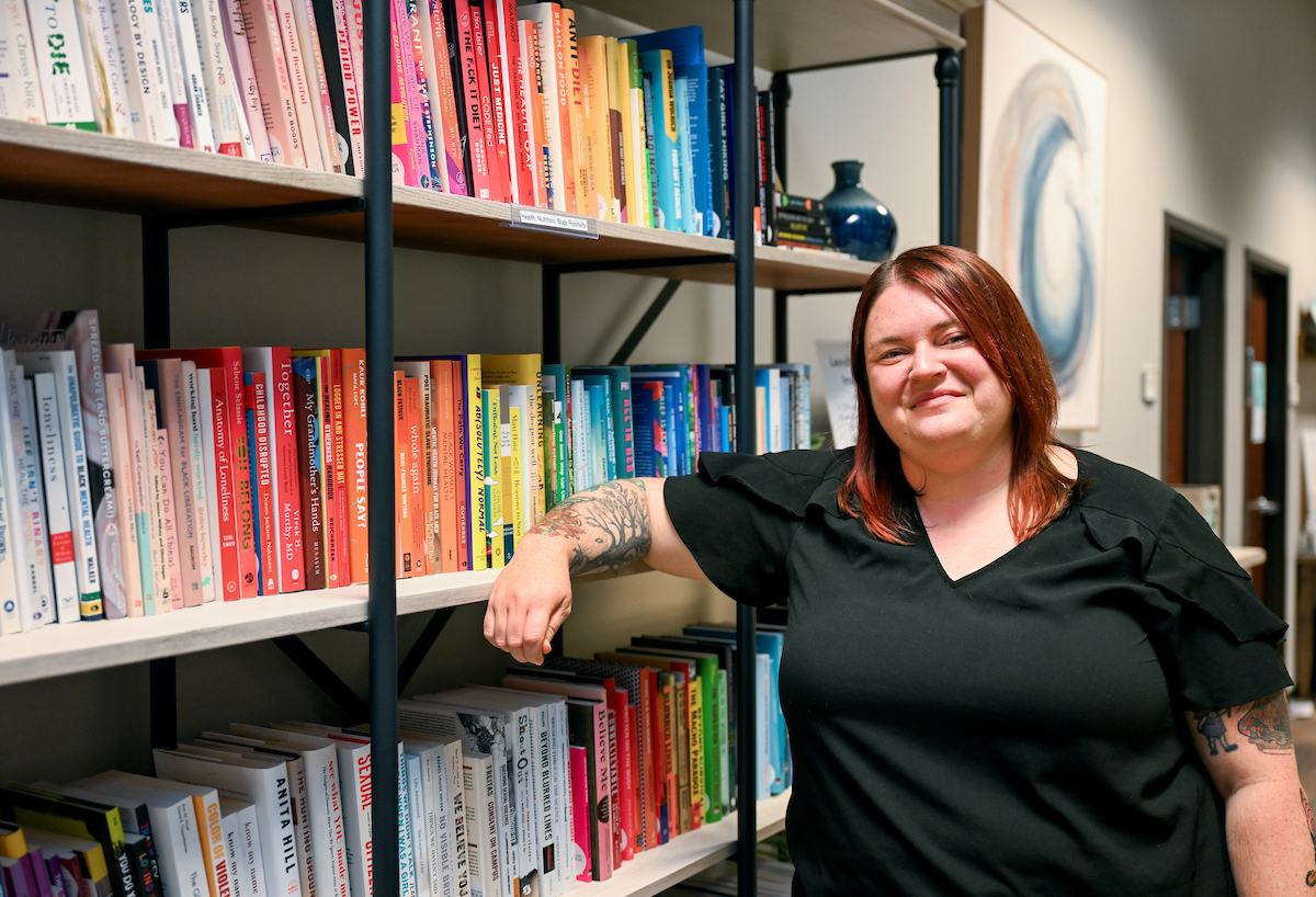 A person with red hair wearing a black blouse leans casually on a bookshelf lined with color-coordinated books. They are smiling at the camera in a hallway with art on the wall and office doors in the background.
