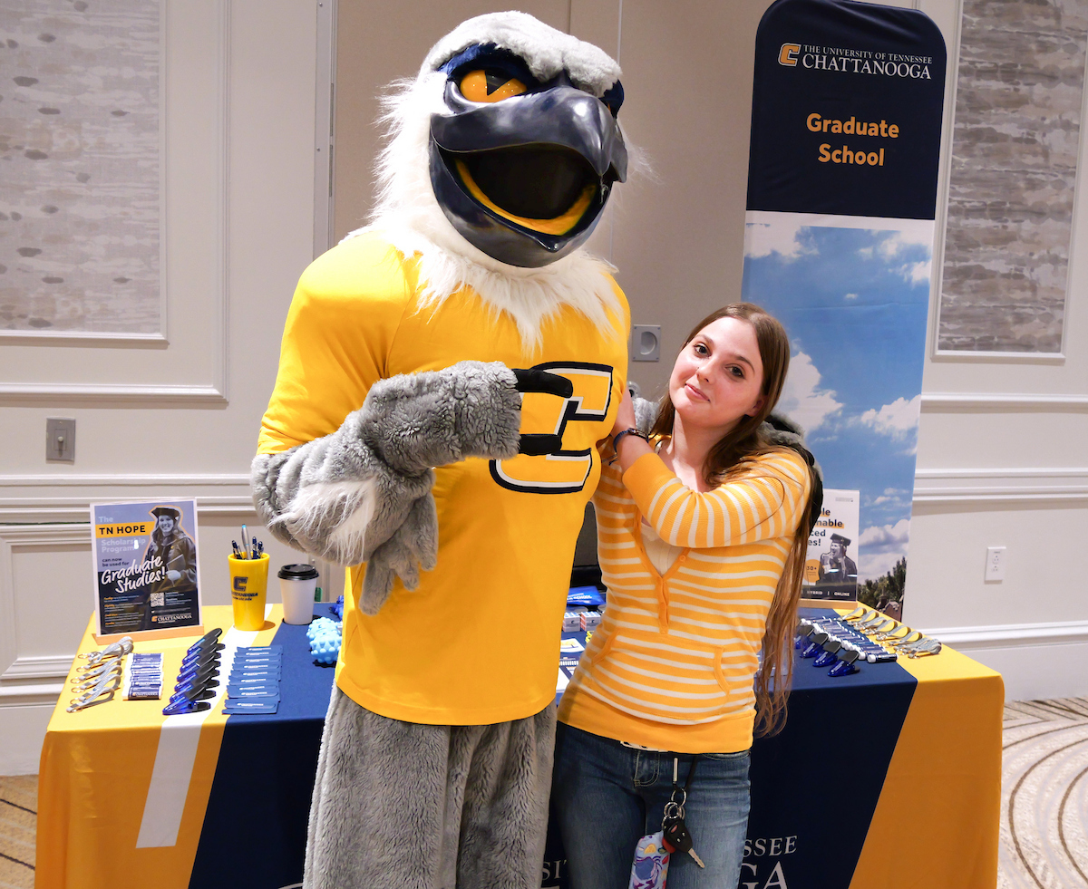 A young woman in a striped yellow shirt poses with mascot, Scrappy. They stand by a UTC table.