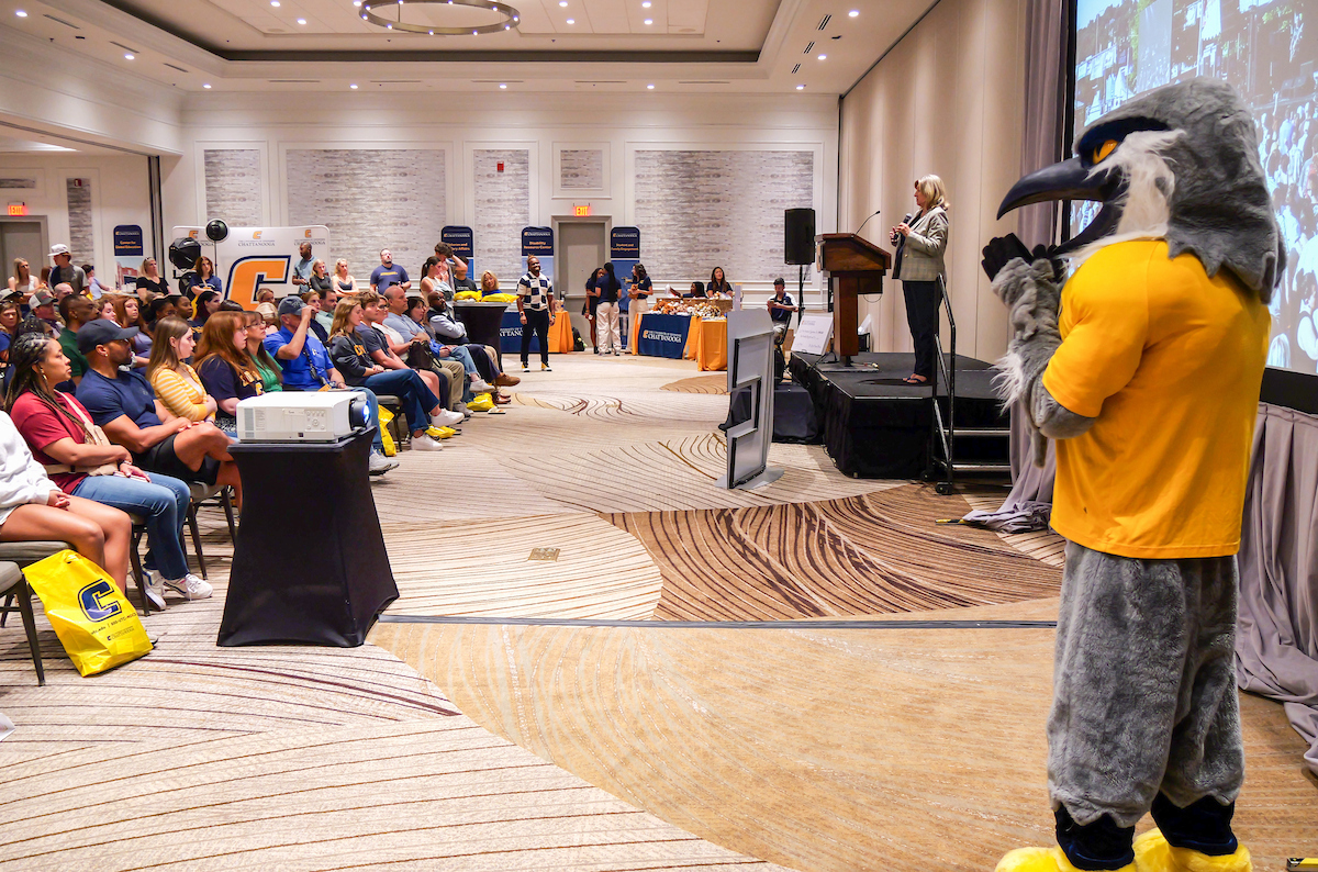 A conference room filled with people listening to a speaker at the podium. Scrappy stands to the side.