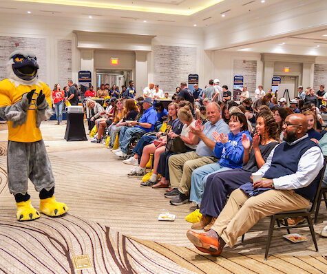 Scrappy stands at the front of a ballroom with hundreds of people at the Power C Tour event