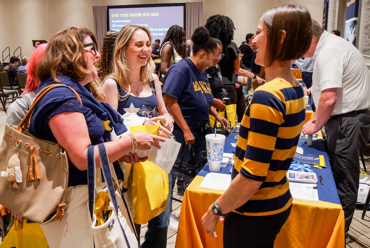 A college fair scene with people chatting at a table. Attendees hold pamphlets and yellow bags.