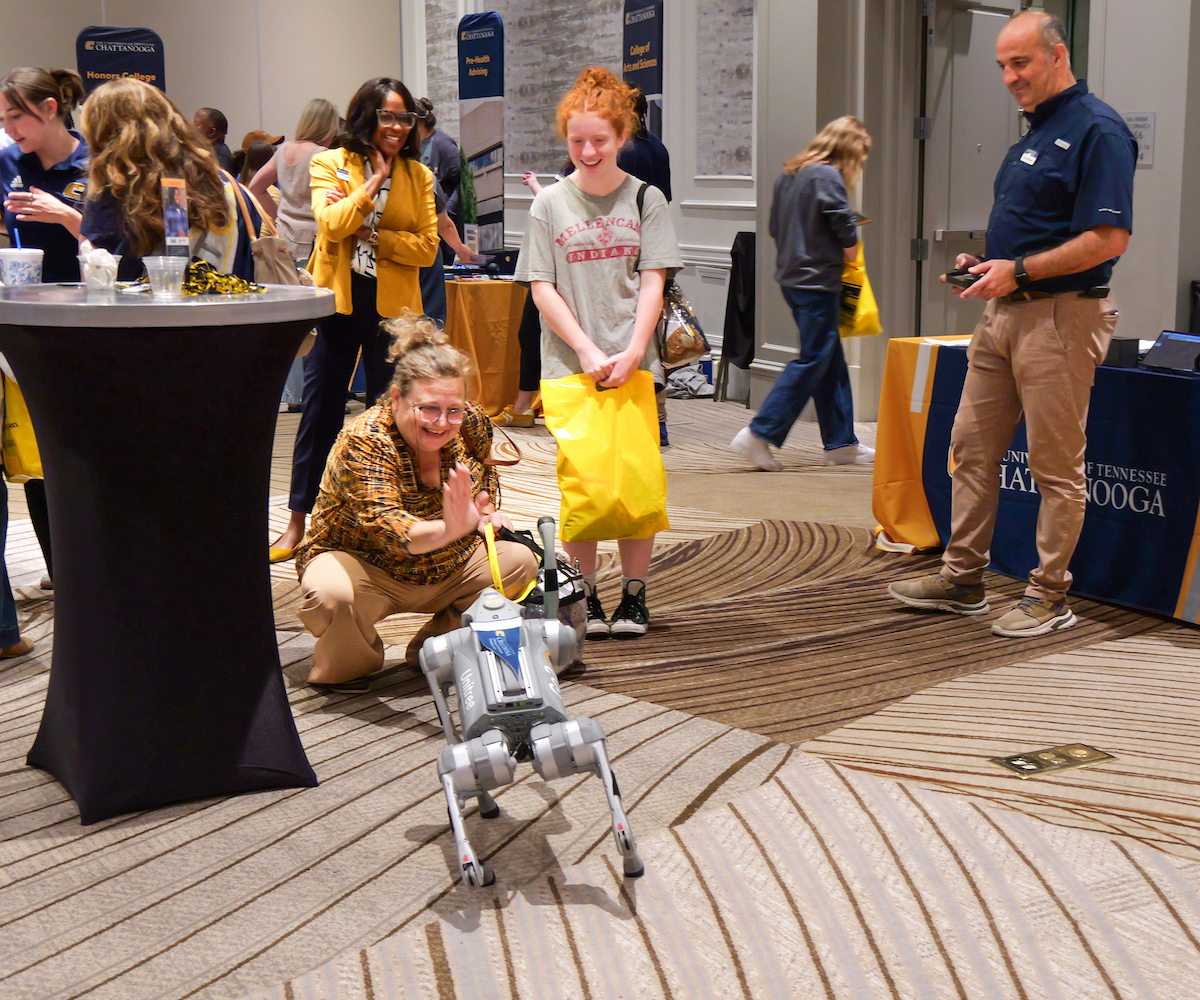 A woman crouches, interacting with a small robot dog, while a smiling young woman stands nearby. People in busy room observe the scene.