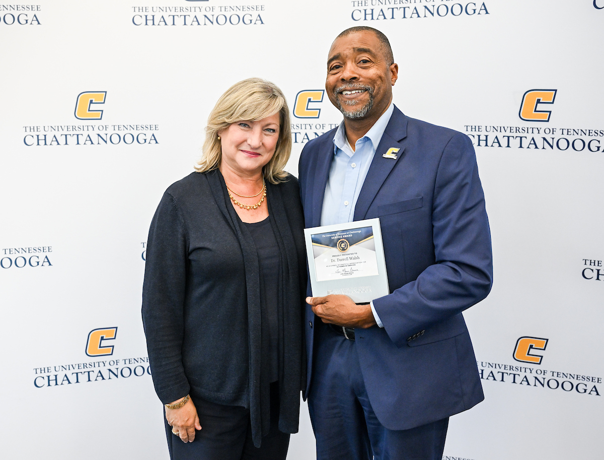 Chancellor Lori Bruce standing next to Dr. Darrell Walsh, who is holding a plaque, in front of a UTC-branded backdrop at the 2025 UTC Service Awards.