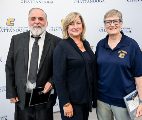 Three people stand together smiling in front of a "University of Tennessee Chattanooga" backdrop. Two hold certificates.