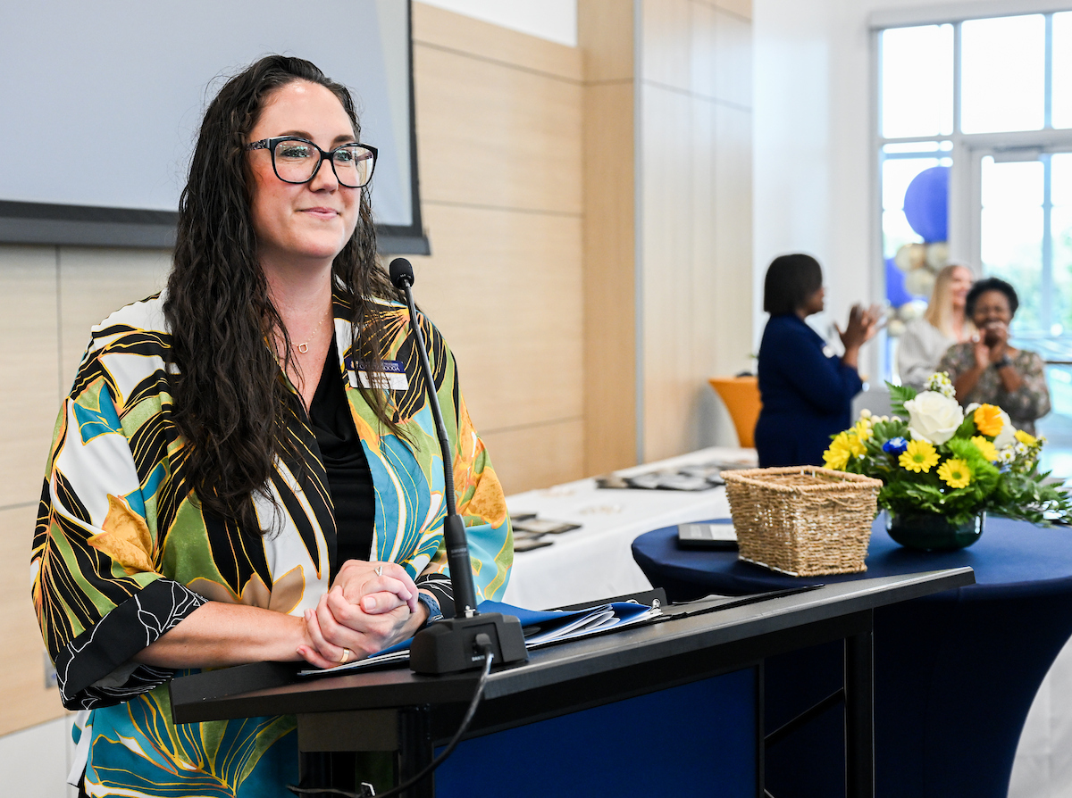 Laure Pou standing at a podium and speaking into a microphone, with a flower arrangement and attendees clapping in the background at the 2025 UTC Service Awards.