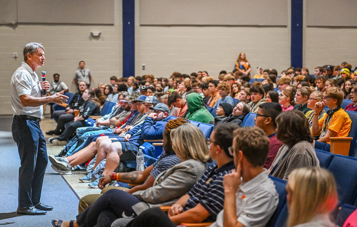 A man speaks into a microphone at the front of a large auditorium filled with students and adults seated in rows of blue chairs. The speaker gestures with one hand while the audience listens, some leaning forward, others taking notes or watching attentively.
