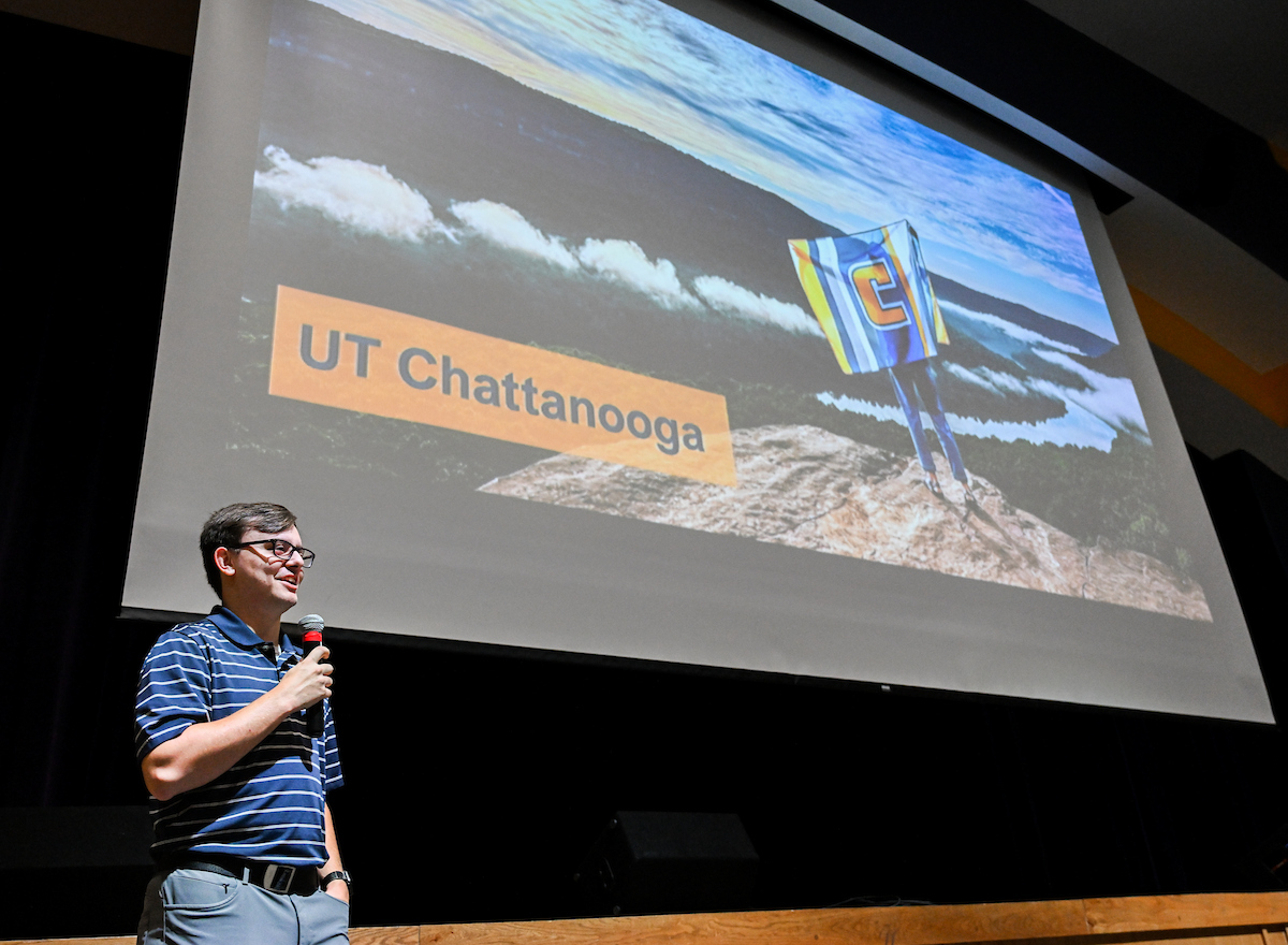 A man holding a microphone stands on stage in front of a large screen displaying an image of someone on a mountaintop holding a UTC flag. The screen includes the text “UT Chattanooga.” The speaker wears glasses, a striped polo shirt, and gray pants.