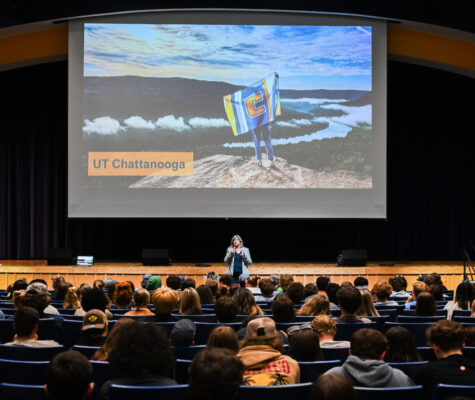 A woman stands on stage speaking to a seated audience in an auditorium. Behind her is a large screen showing a scenic mountaintop view with a person holding a UTC flag and the words “UT Chattanooga.”