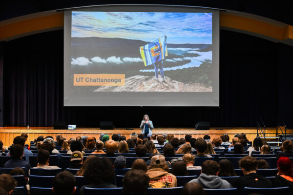 A woman stands on stage speaking to a seated audience in an auditorium. Behind her is a large screen showing a scenic mountaintop view with a person holding a UTC flag and the words “UT Chattanooga.”
