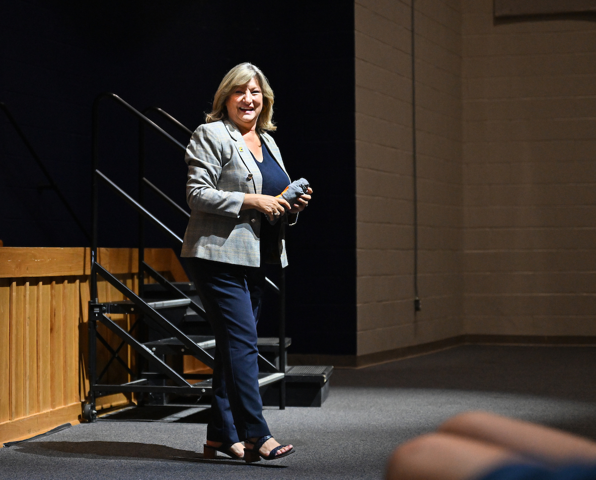 A woman stands and smiles while holding a small rolled item, speaking to an unseen audience in an auditorium. She wears a gray plaid blazer over a navy top and pants with black sandals. A wooden stage, black railings, and a brick wall are in the background.