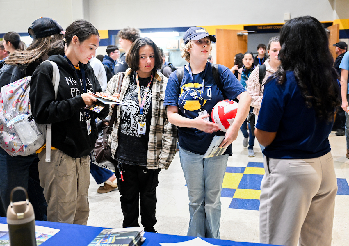 Three students talk with a representative at an information table covered with brochures and flyers. One student holds a volleyball and another flips through printed materials while others look on. The room is busy with more people in the background.