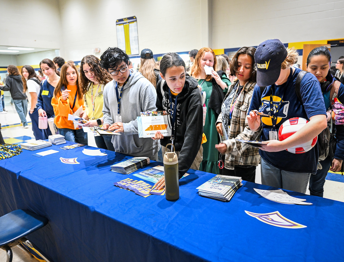 A group of students lines up at a blue-covered information table filled with UTC brochures, stickers, and handouts. Some pick up materials and others read pamphlets. The area is busy with many students behind them.