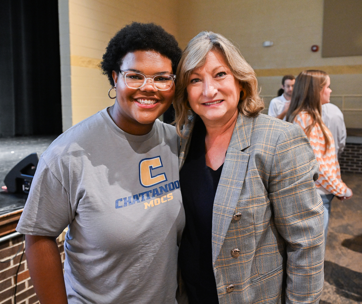 Two women smile for a photo inside an auditorium. One wears a gray “Chattanooga Mocs” T-shirt and glasses; the other wears a gray plaid blazer over a black shirt. People and a stage are visible in the background.
