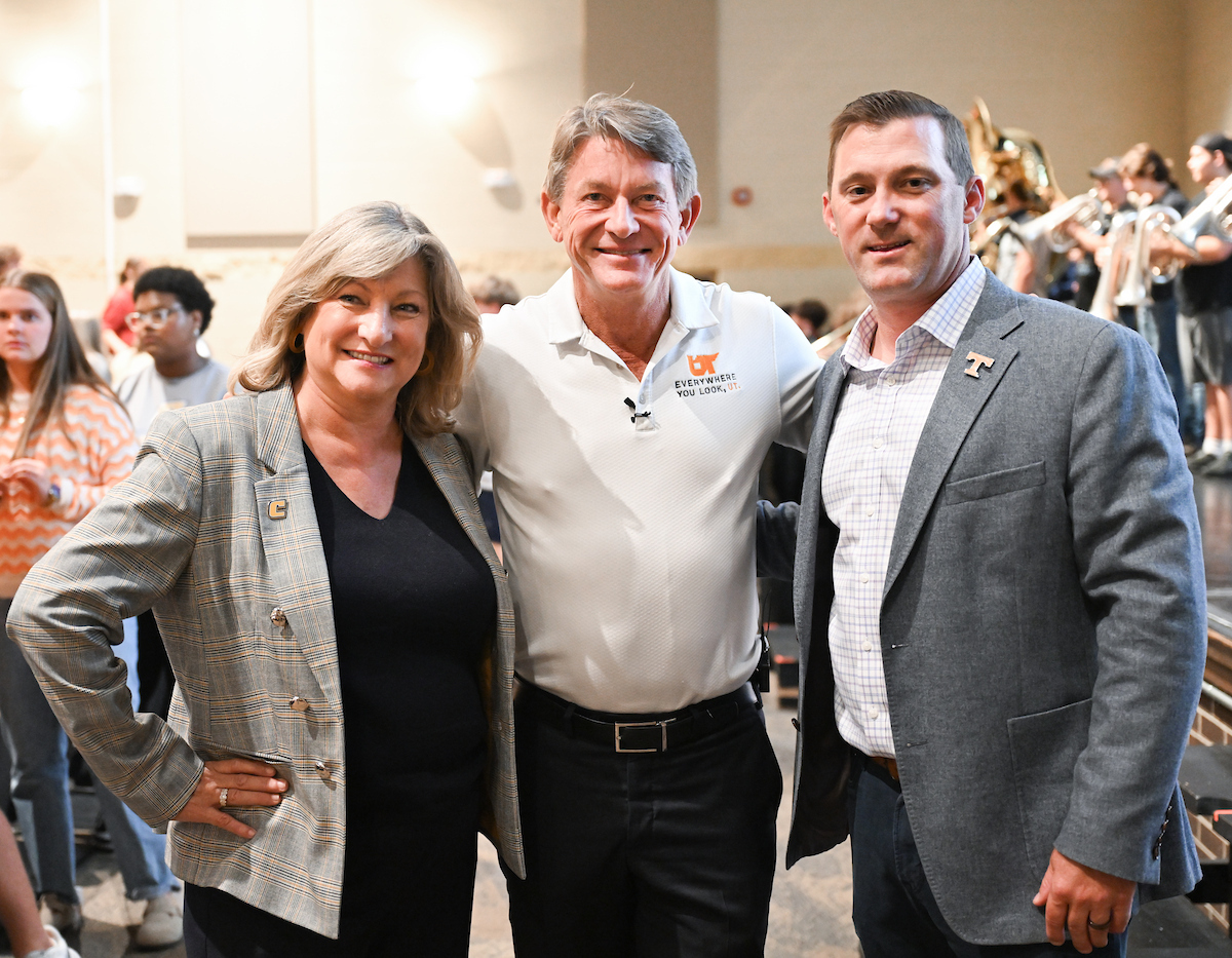 Three adults stand together smiling for a photo in an auditorium. The woman on the left wears a gray plaid blazer, the man in the center wears a white polo shirt, and the man on the right wears a gray blazer.