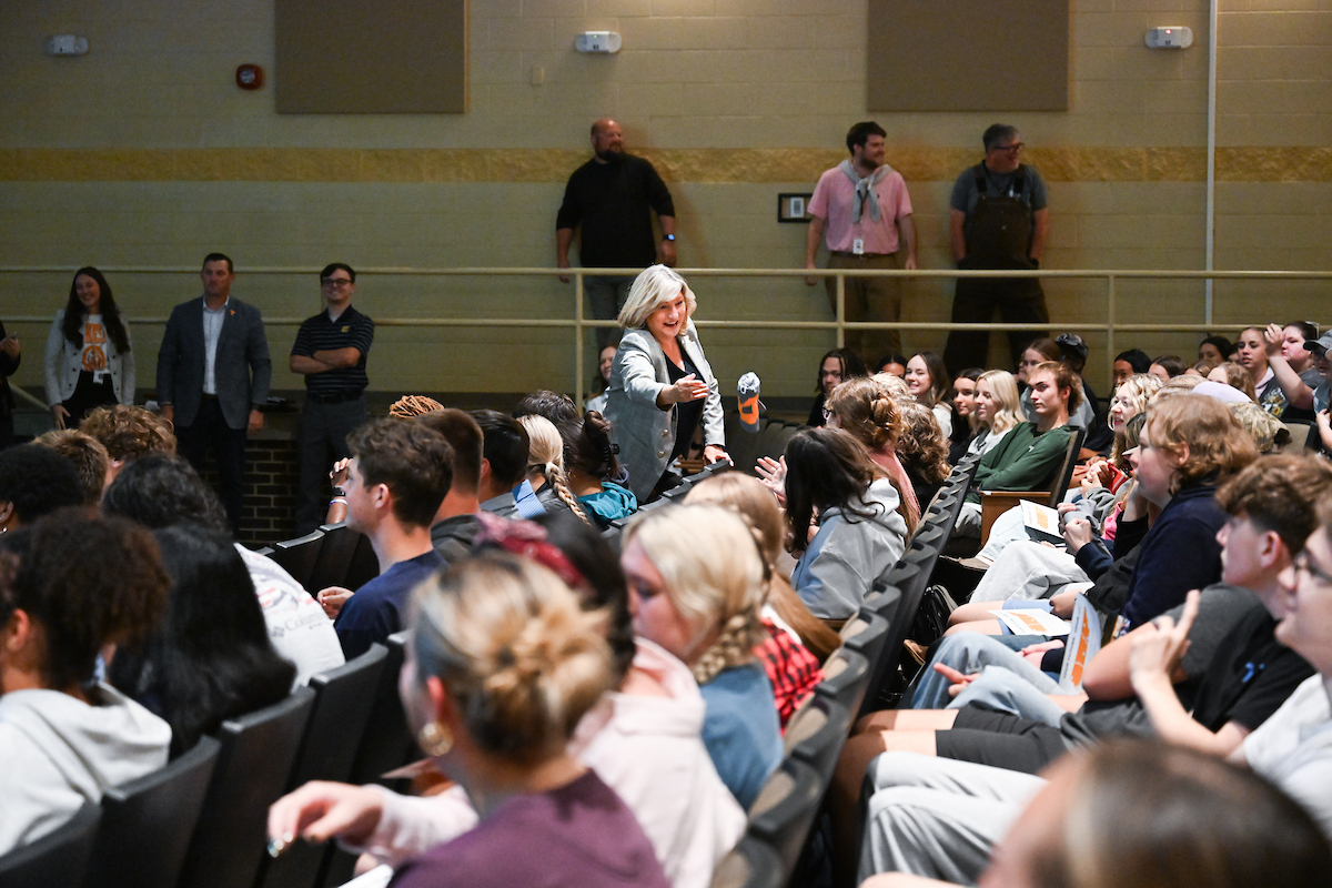 A woman stands in an auditorium aisle handing a t shirt to a student while smiling. Rows of seated students watch and some reach out their hands. Several adults stand along the back wall.