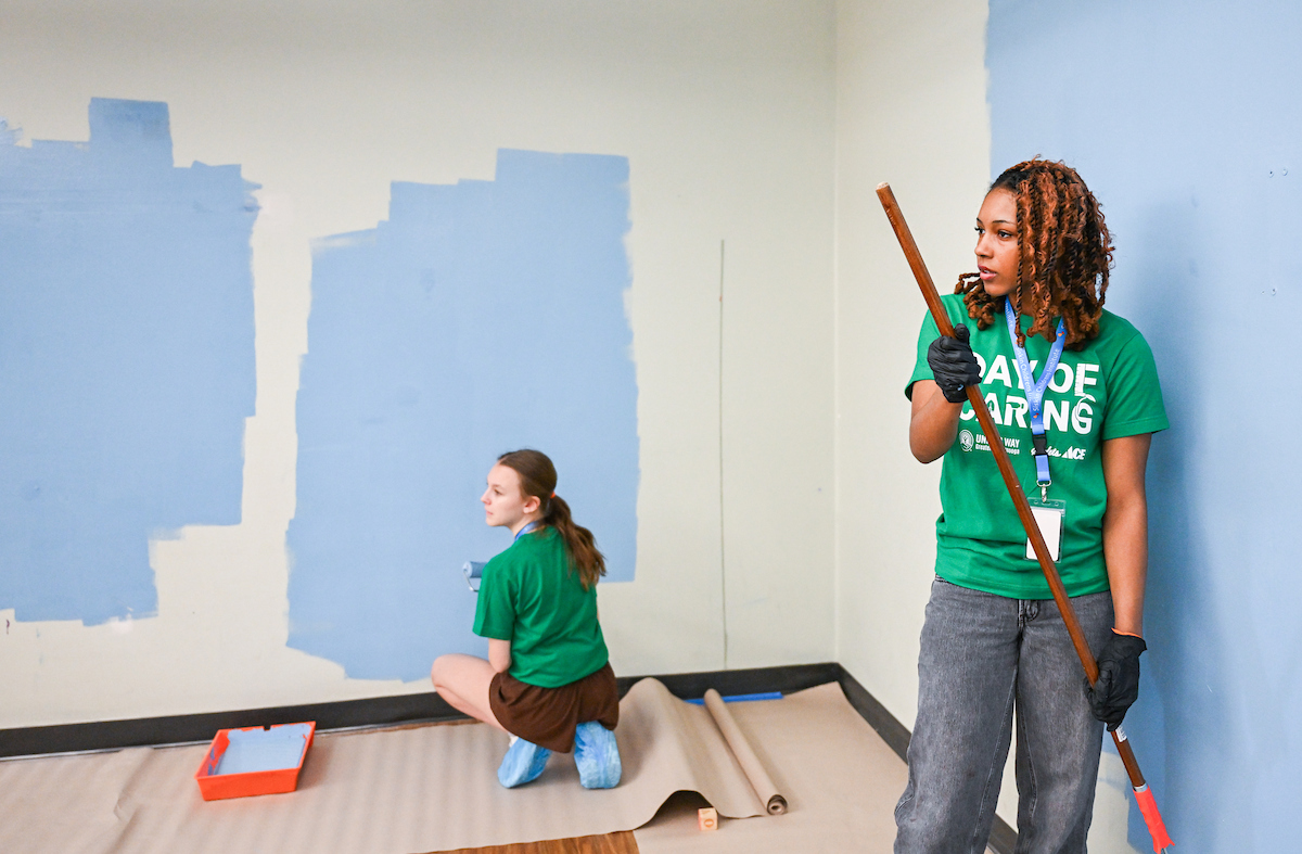 Two young women hold roller paint brushes and paint a light blue wall
