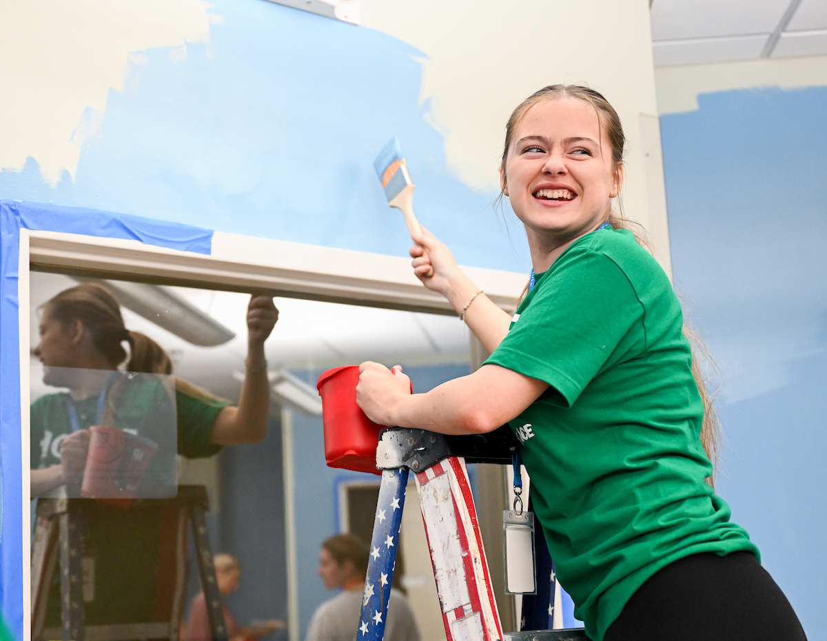 Young woman wearing a green shirt smiles on a ladder as she paints a light blue wall.