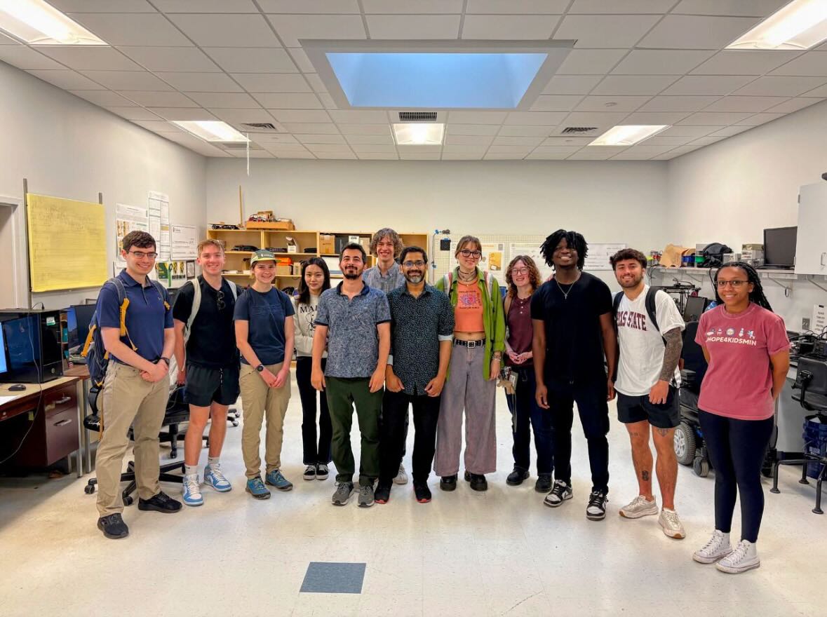 A group of 12 people stands smiling in a bright lab or workshop space with computers and equipment on the sides. Most are casually dressed, some wearing backpacks. Shelves and posters with diagrams and notes are visible in the background.