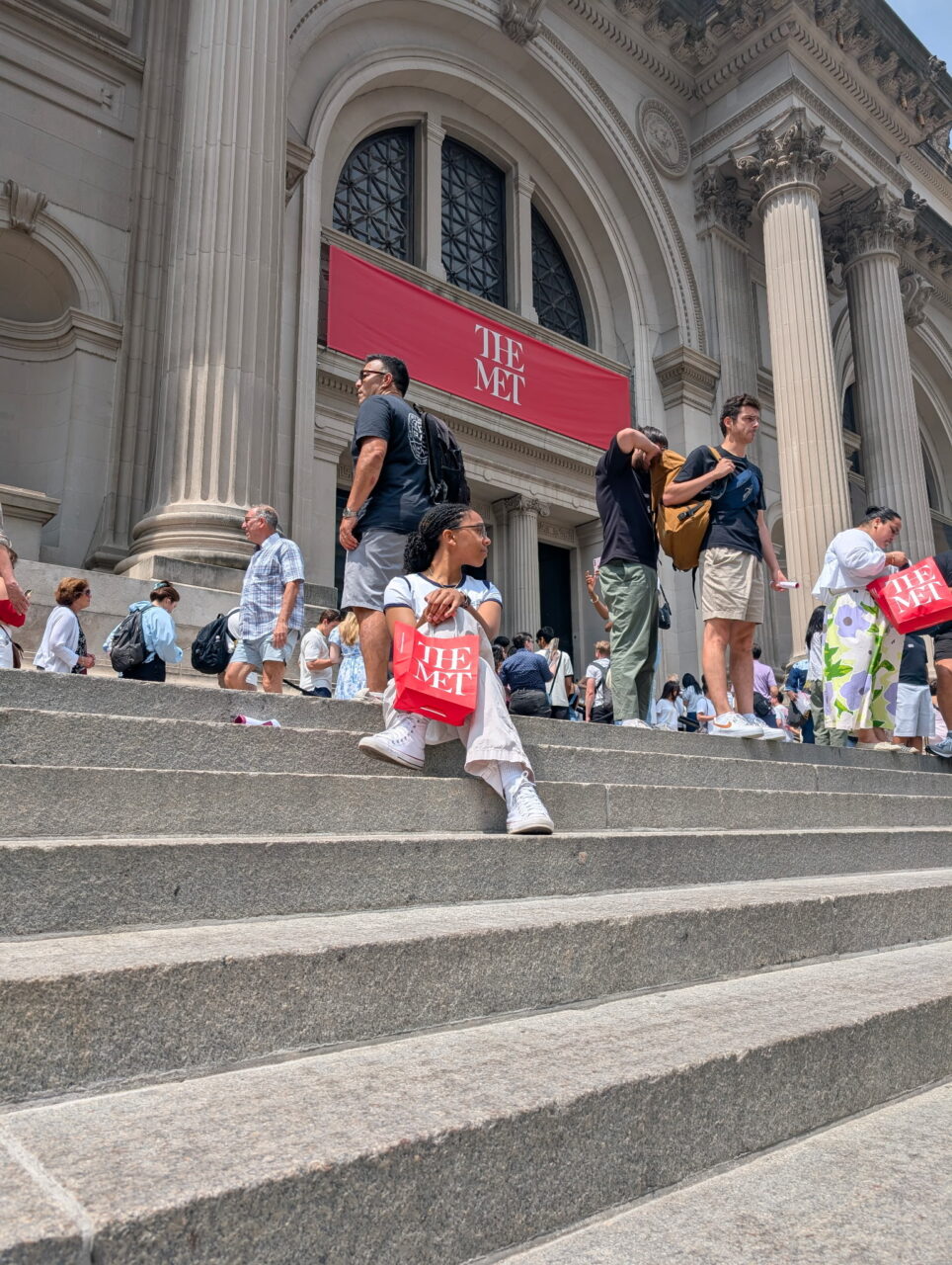 Student Hailey Henderson sits on the wide stone steps outside the Metropolitan Museum of Art in New York City, holding a red museum shopping bag with “THE MET” printed on it. Other visitors are standing and walking nearby, with the museum’s grand columns and a red sign reading “THE MET” in the background.
