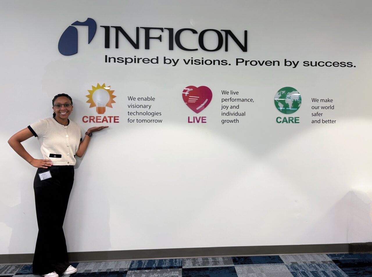Student Hailey Henderson stands indoors, smiling and gesturing toward a colorful corporate wall display for INFICON with the words “Inspired by visions. Proven by success.” and icons labeled CREATE, LIVE and CARE with short taglines about technology, growth, and safety.