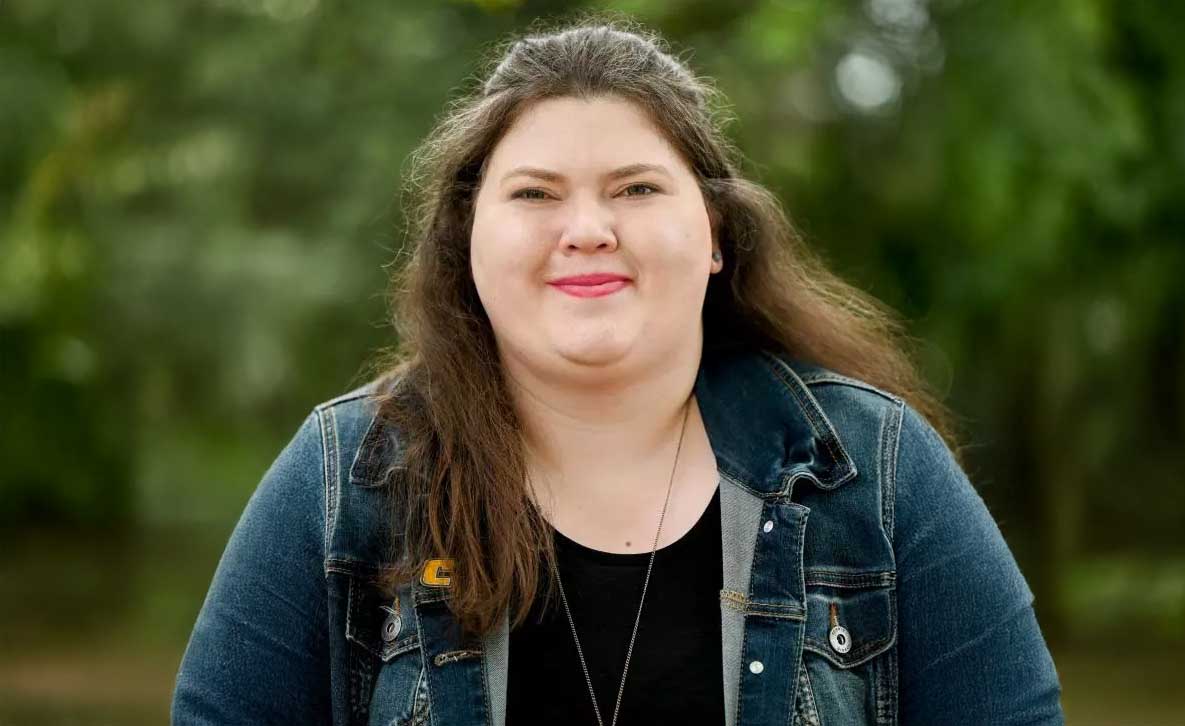 A person with long brown hair smiles softly while standing outdoors in front of blurred greenery. They are wearing a denim jacket over a black top with a necklace and a small gold “C” pin on the jacket.