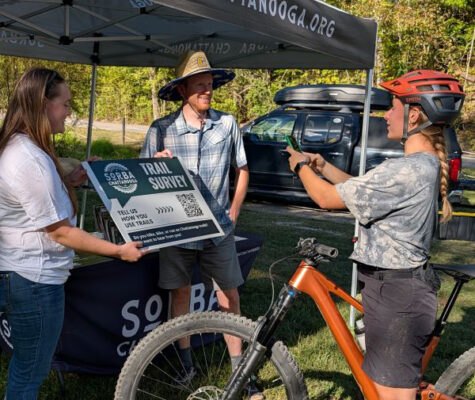 Student Chloe Wesson and Dr. Ryan Zwart hold a sign saying Trail Survey while a bicyclist scans the sign's QR code