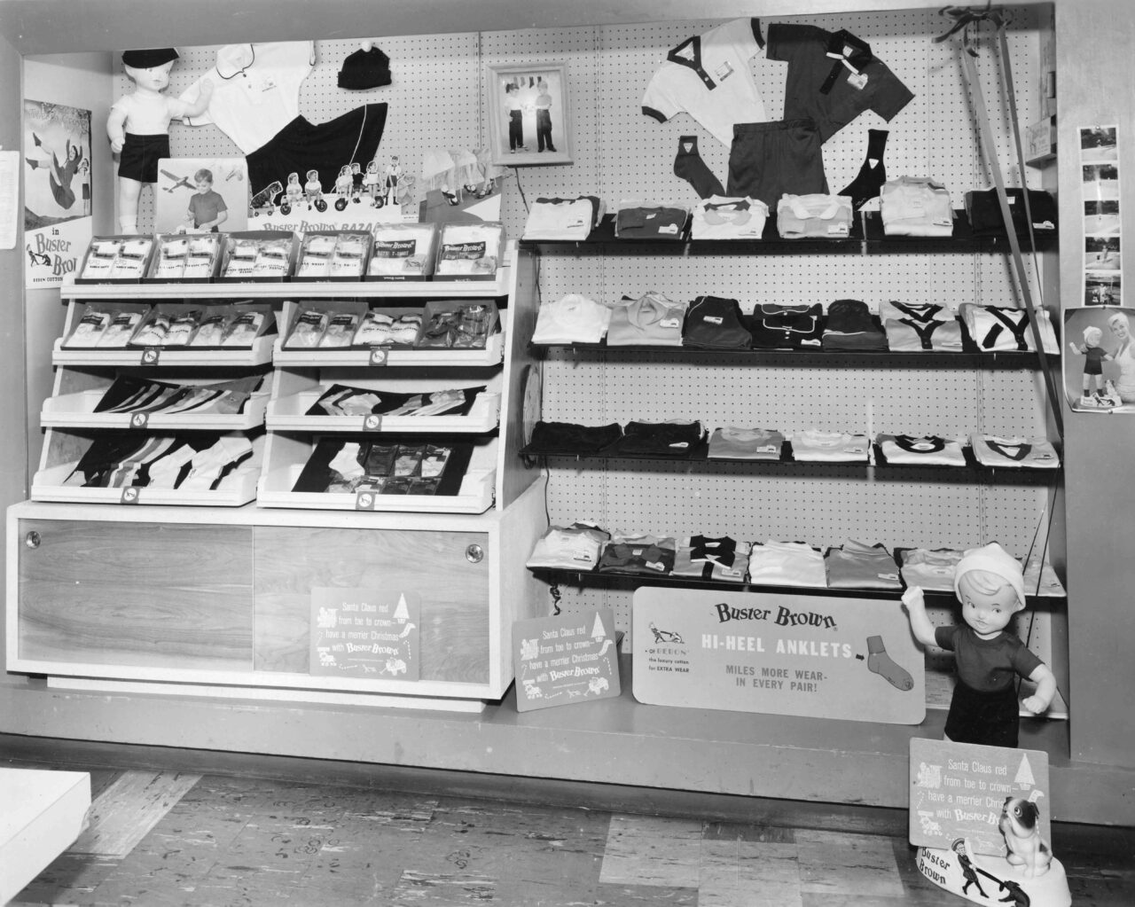 A black-and-white photo of a Buster Brown retail display shows shelves filled with neatly arranged children’s socks, shirts, and folded clothing. The display features Buster Brown branding, including signs advertising “Hi-Heel Anklets” and several cutouts of the cartoon character. A Buster Brown mannequin waves from the right side of the image, while posters and photographs decorate the pegboard wall behind the merchandise.