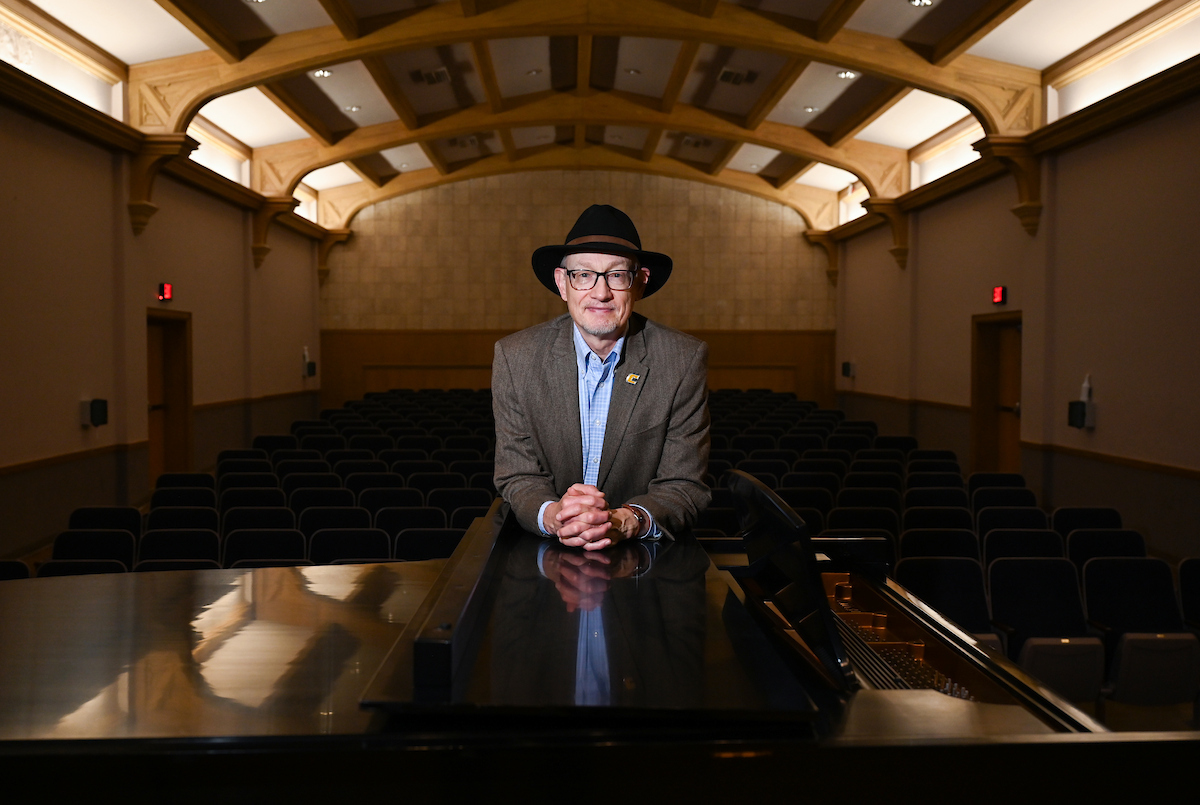 Jonathan McNair, wearing a dark hat, glasses and a brown jacket over a light blue shirt, leans on a grand piano in an empty performance hall. Rows of black seats and the hall’s wooden ceiling beams frame the background as warm lighting highlights McNair at center.