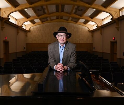 Jonathan McNair, wearing a dark hat, glasses and a brown jacket over a light blue shirt, leans on a grand piano in an empty performance hall. Rows of black seats and the hall’s wooden ceiling beams frame the background as warm lighting highlights McNair at center.