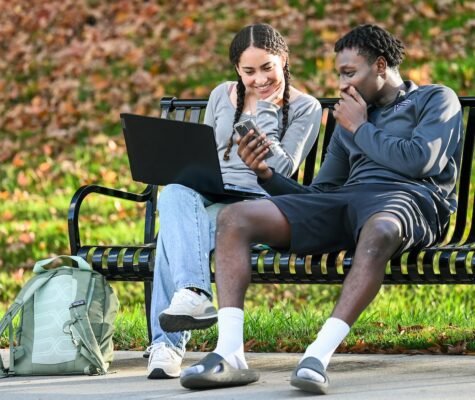 Two students sit on a bench sharing a phone screen, smiling.
