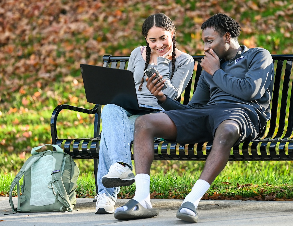 Two college students sitting together on a black metal bench outdoors on a sunny fall day. The student on the left, smiling, holds a laptop on their lap while leaning toward the student on the right, who is showing something on a phone. A green backpack rests on the ground near their feet, and fallen leaves cover the grassy background.